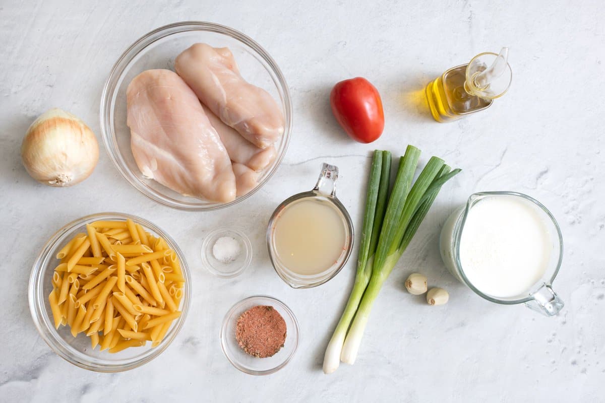 Ingredients for recipe before being prepped: onion, uncooked pasta, raw chicken breast, seasonings, broth, green onions, roma tomato, oil, garlic cloves, and cream.