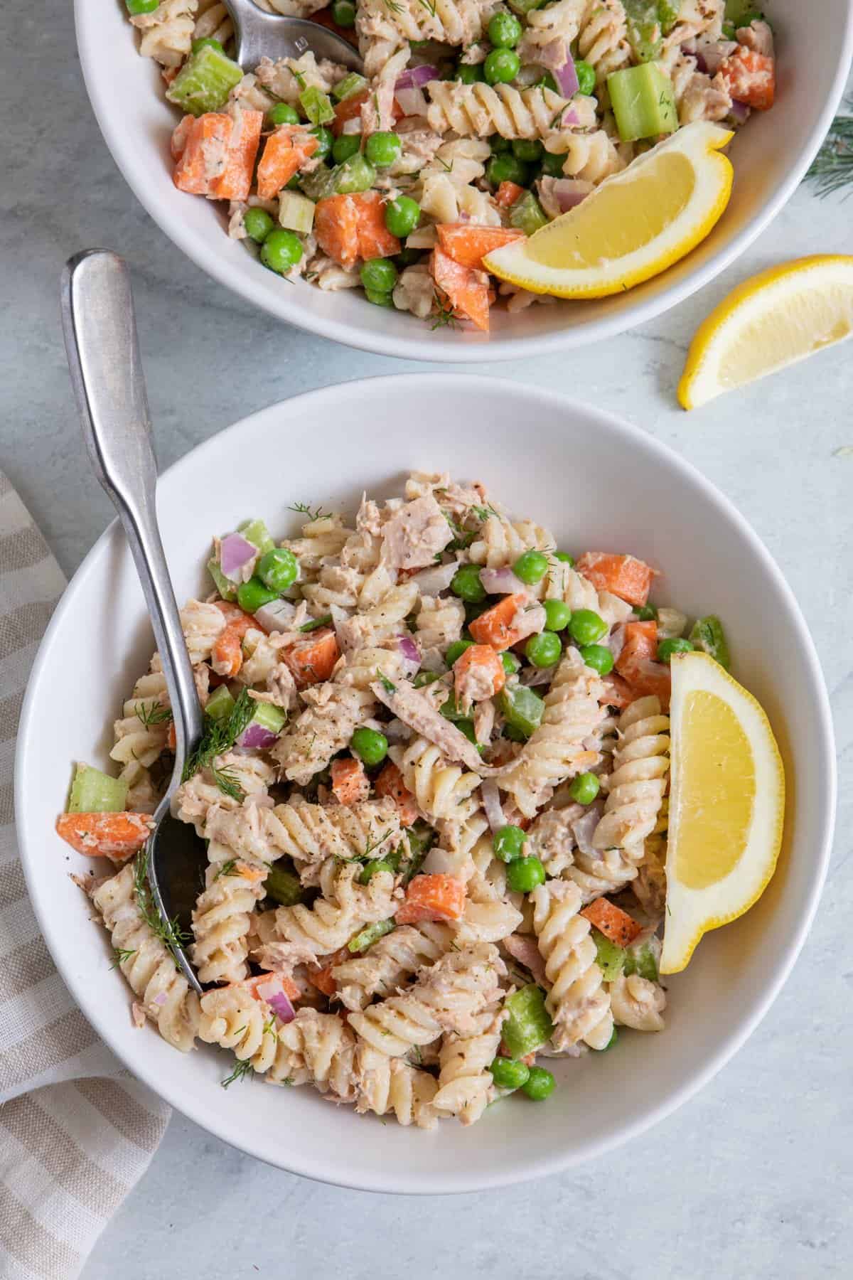 Two small bowls of tuna pasta salad with spoon and one bowl off shot, garnished with lemon wedges.