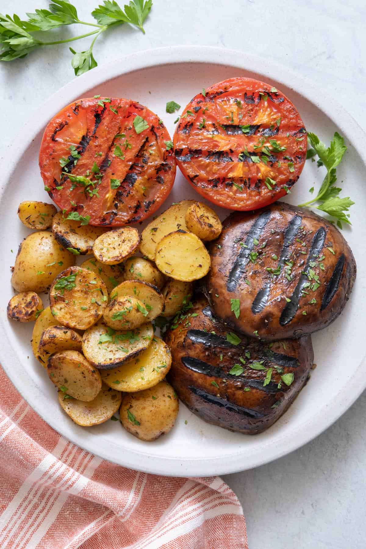 2 grilled portabellas, 2 halved grilled tomatoes, and grilled halved baby potatoes on a plate garnished with parsley.