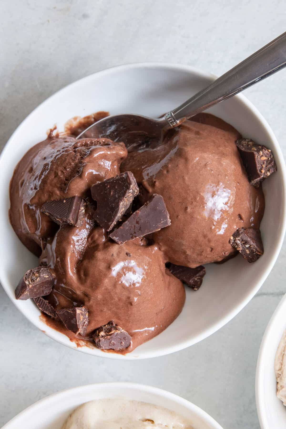 Chocolate Nice Cream in white bowl with chunks of chocolate added and a spoon dipped into the side.