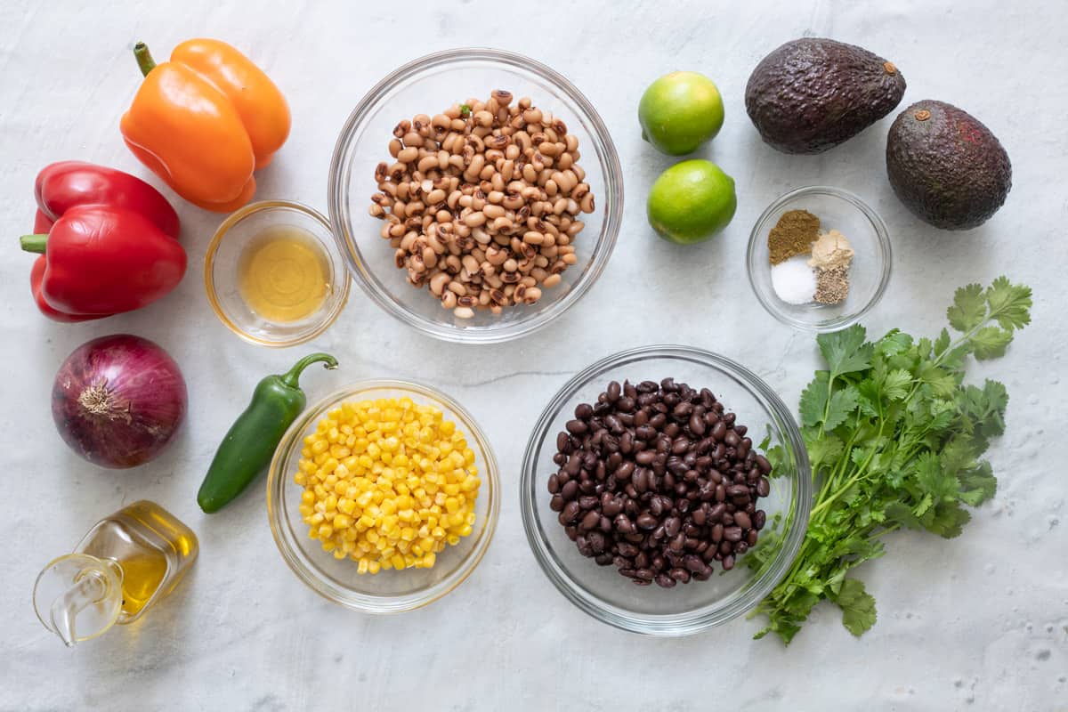 Ingredients for recipe before prepped: red bell pepper, orange bell pepper, red onion, oil, jalapeno pepper, honey, corn, black eye peas, black beans, 2 limes, 2 avocados, seasonings, and cilantro.