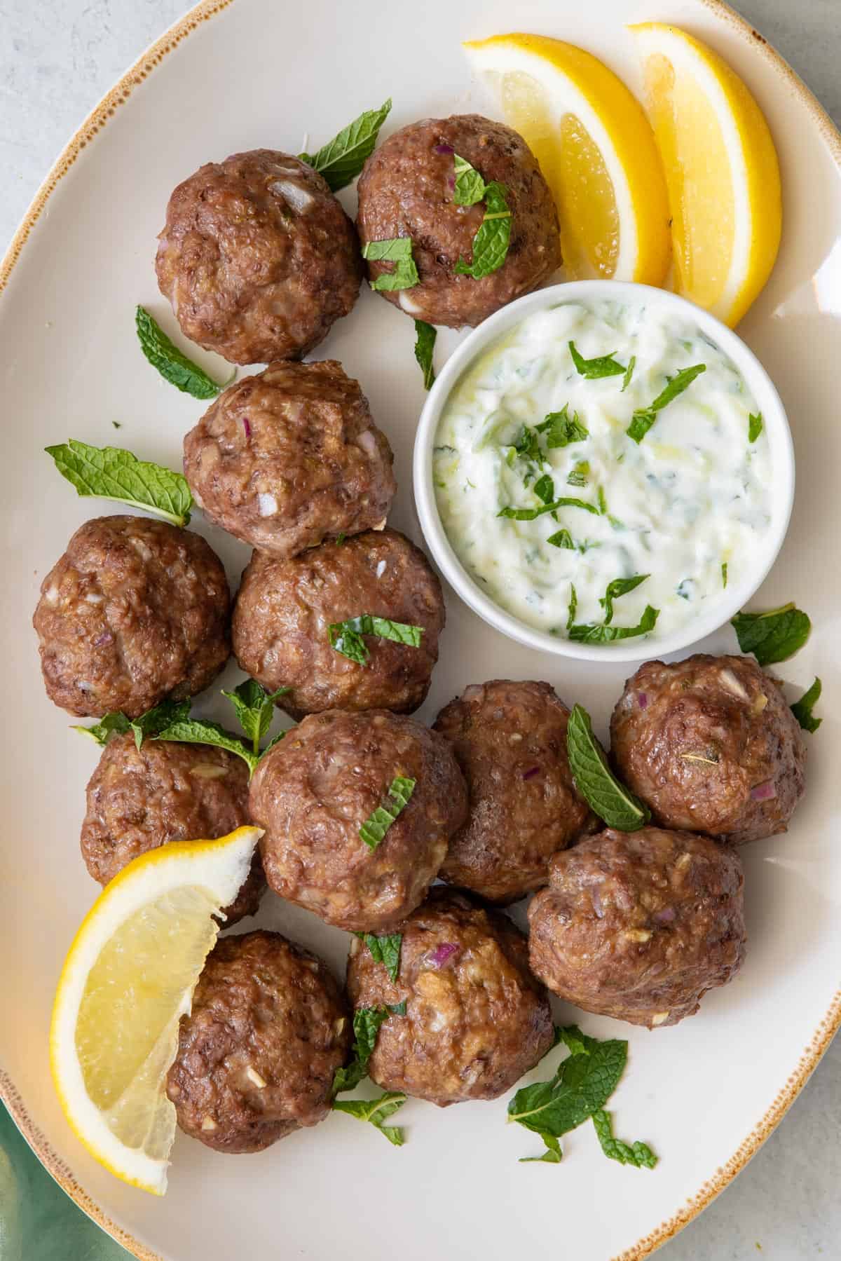 Oval platter with meatballs and side of tzatziki sauce in a small bowl. Garnished with lemon wedges and fresh mint leaves.