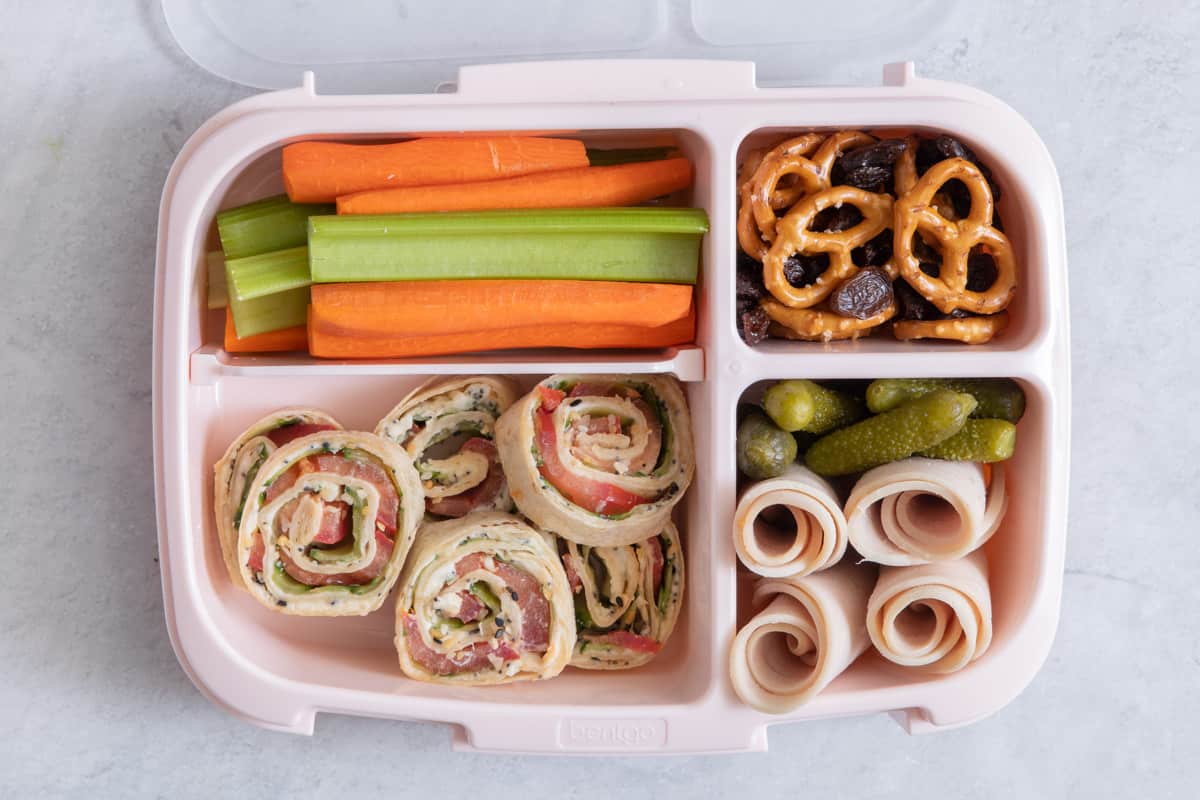 Lunchbox with individual sections with different lunch foods for kids: celery and carrot sticks, pretzels with raisins, turkey roll ups, and everything bagel pinwheels.