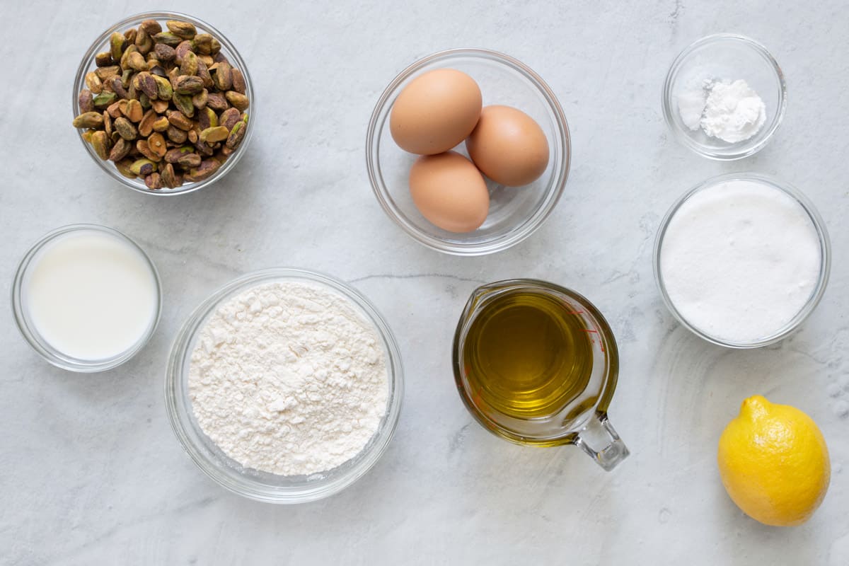 Ingredients for bread recipe before being prepped in individual bowls: roasted pistachios, milk, flour, 3 eggs, oil, baking powder and salt, sugar, and a whole lemon.