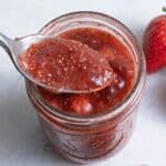 Strawberry chia jam in a jar with a spoon holding up some and strawberries nearby.