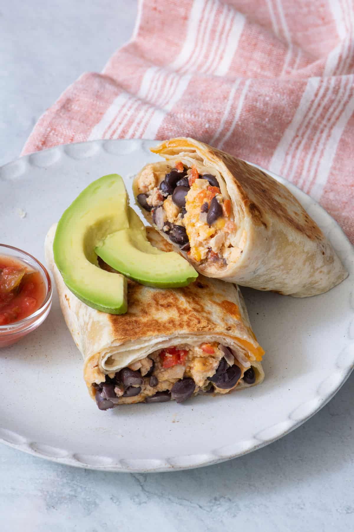 Burrito cut in half on a plate with slices of avocado on top and a small side of salsa.