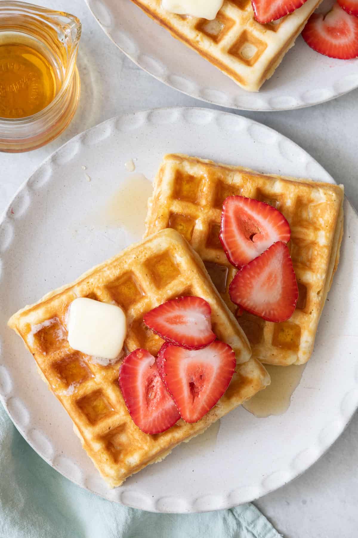 Overhead shot of a plate of square waffles with strawberry slices, melted butter, and syrup and another plate sitting off frame next to a carafe of maple syrup.