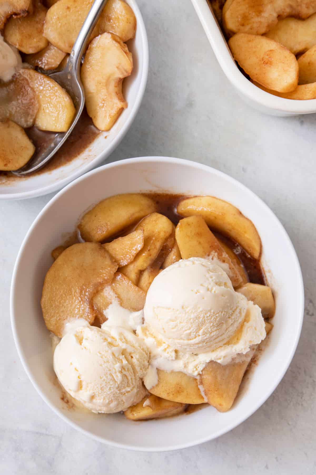 Small bowl of baked apple slices with ice cream scooped on top and extra apples to the side.