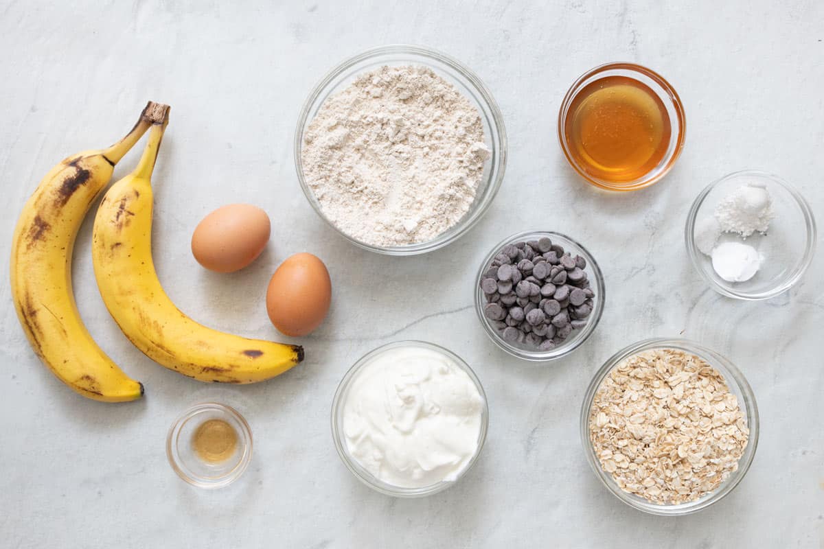 Ingredients for muffin recipe: 2 ripe bananas, 2 eggs, vanilla, oat flour, Greek yogurt, chocolate chips, honey, baking powder, baking soda, salt, and quick oats.