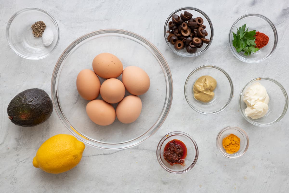 Ingredients for 3 different flavors before being prepped: salt and pepper, avocado, lemon, 5 eggs in a bowl, black olives, mustard, harissa maste, spices, mayo, and turmeric.
