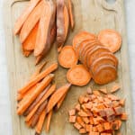 A cutting board with sweet potatoes cut 4 different ways: wedges, slices, sticks, and cubes.