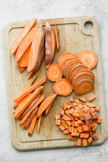 A cutting board with sweet potatoes cut 4 different ways: wedges, slices, sticks, and cubes.