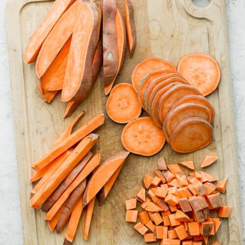 A cutting board with sweet potatoes cut 4 different ways: wedges, slices, sticks, and cubes.