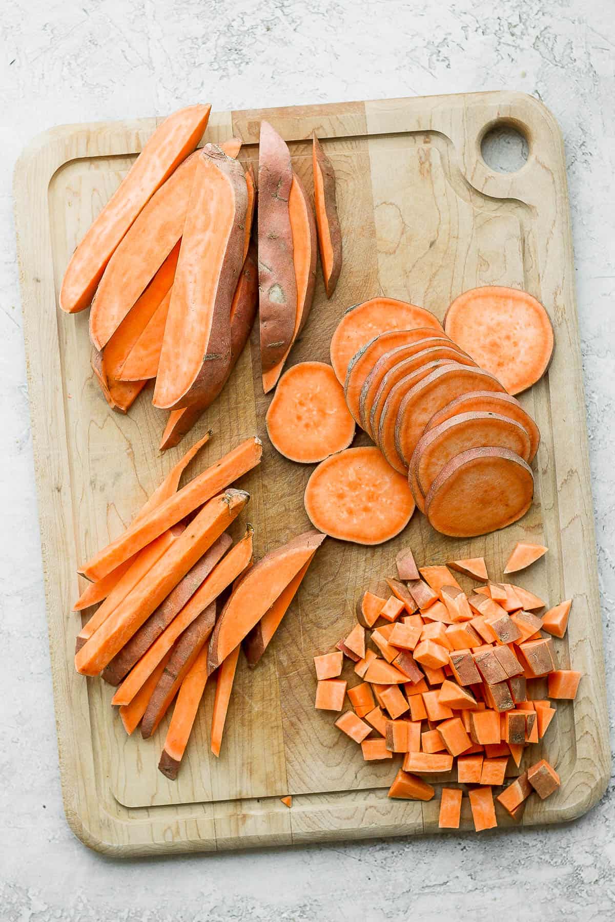 A cutting board with sweet potatoes cut 4 different ways: wedges, slices, sticks, and cubes.