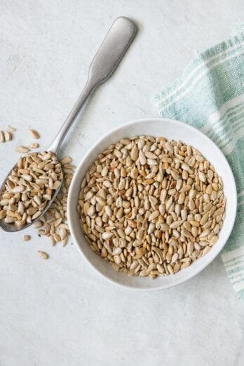 Small bowl of roasted sunflower seeds with spoon next to bowl overflowing with seeds.