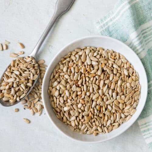 Small bowl of roasted sunflower seeds with spoon next to bowl overflowing with seeds.