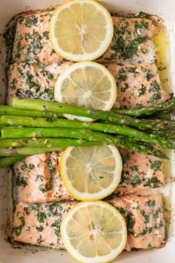 Roasted salmon and asparagus in a baking dish with lemons resting over it.