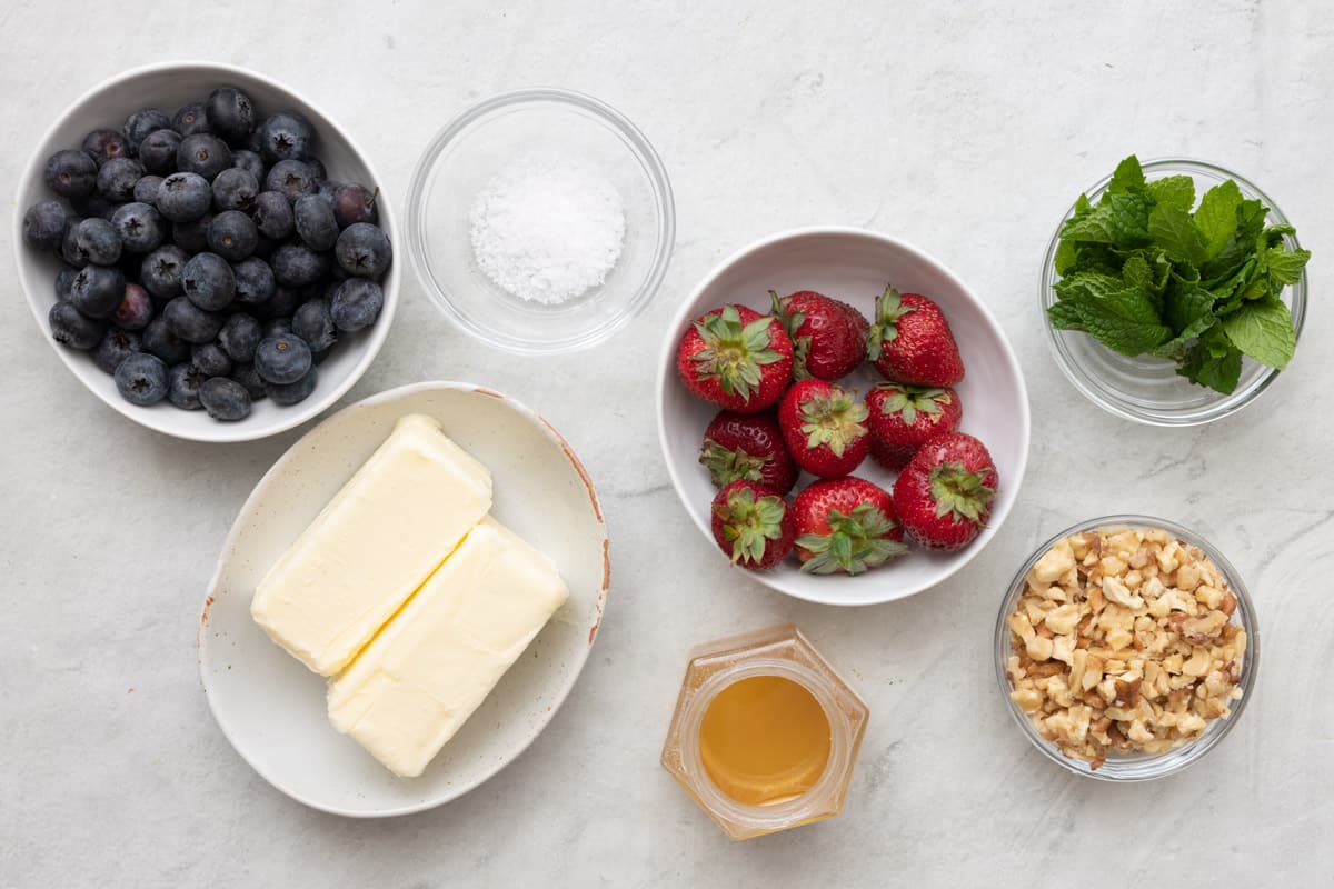 Ingredients for recipe in individual bowls: fresh blueberries, butter sticks, salt, strawberries, mint leaves, honey, and walnuts.