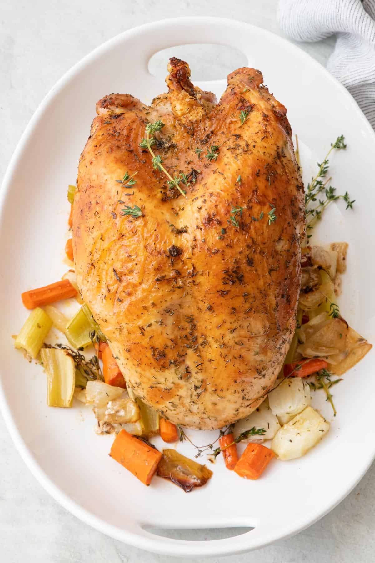 Whole roasted turkey breast on top of roasted veggies garnished with fresh herbs on a large oval platter.