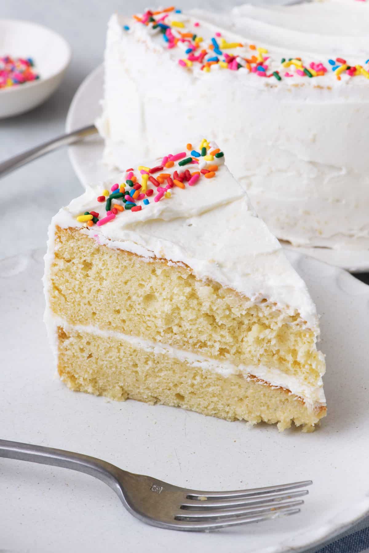 Slice of 2 layer white cake with frosting and sprinkles on a small plate with a fork and whole cake behind it.