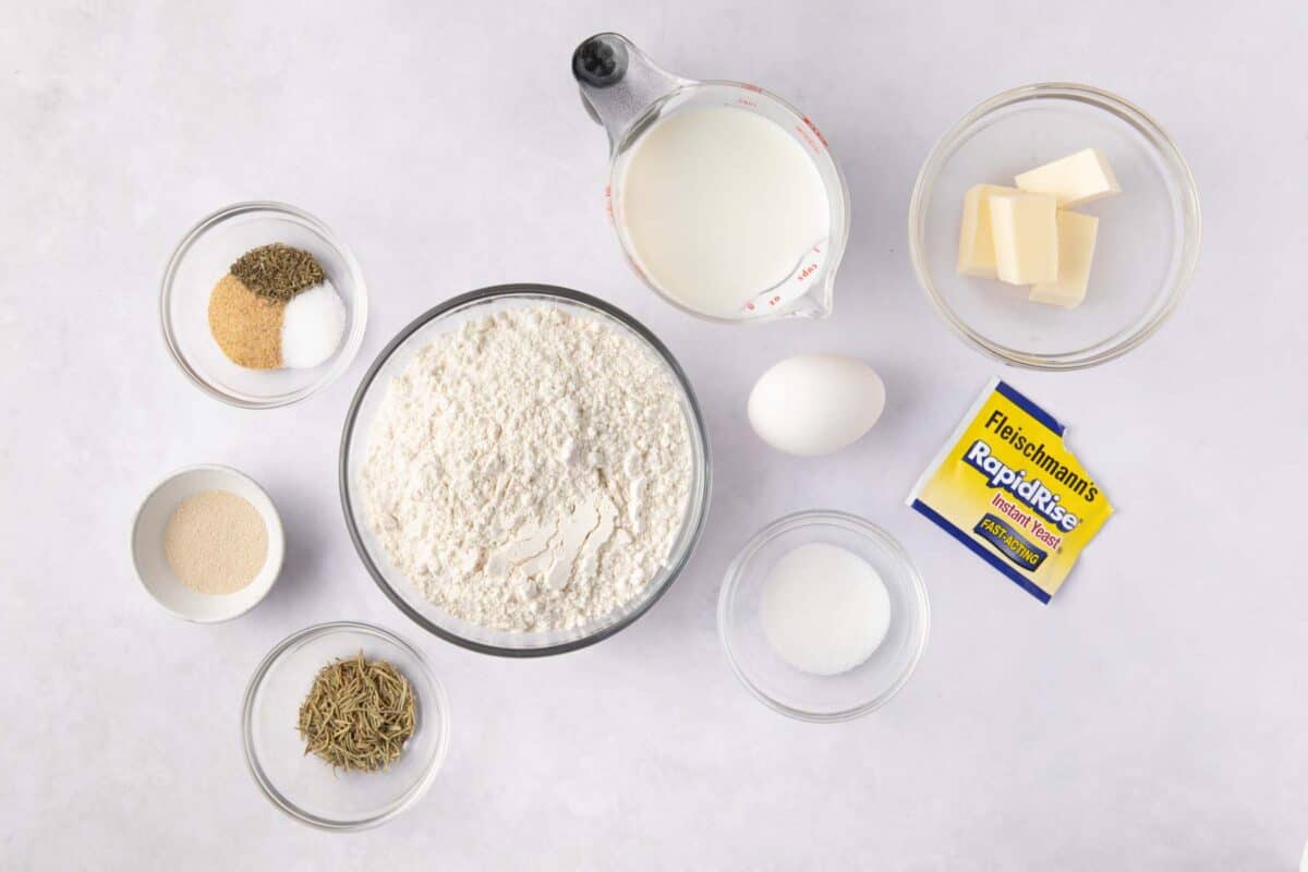 Ingredients for recipe in individual bowls: garlic powder, salt, and rosemary, thyme, sugar, flour, milk, butter, an egg, and Fleischmannโsยฎ RapidRiseยฎ Instant Yeast package with contents in a small bowl.