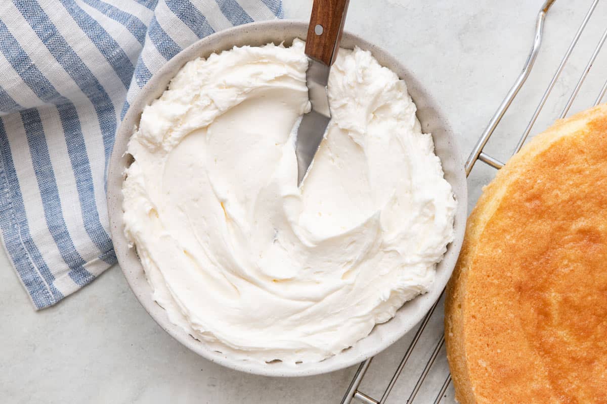 Bowl of frosting with cake spatula dipped in and cake on cooling rack next to it.