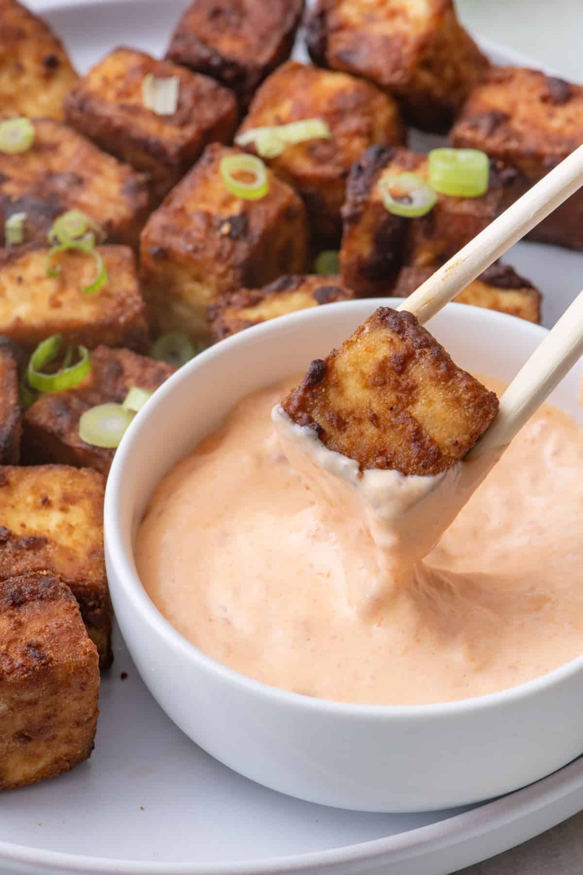 Chopsticks picking up air fryer tofu from platter with a small bowl of spicy mayo.