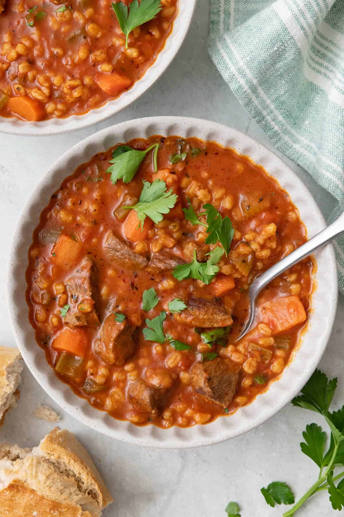 Bowl of barley and beef soup garnished with parsley and a spoon dipped in with bread nearyby.