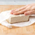 Hand firmly pressing tofu that is wrapped in paper towels on a cutting baord.