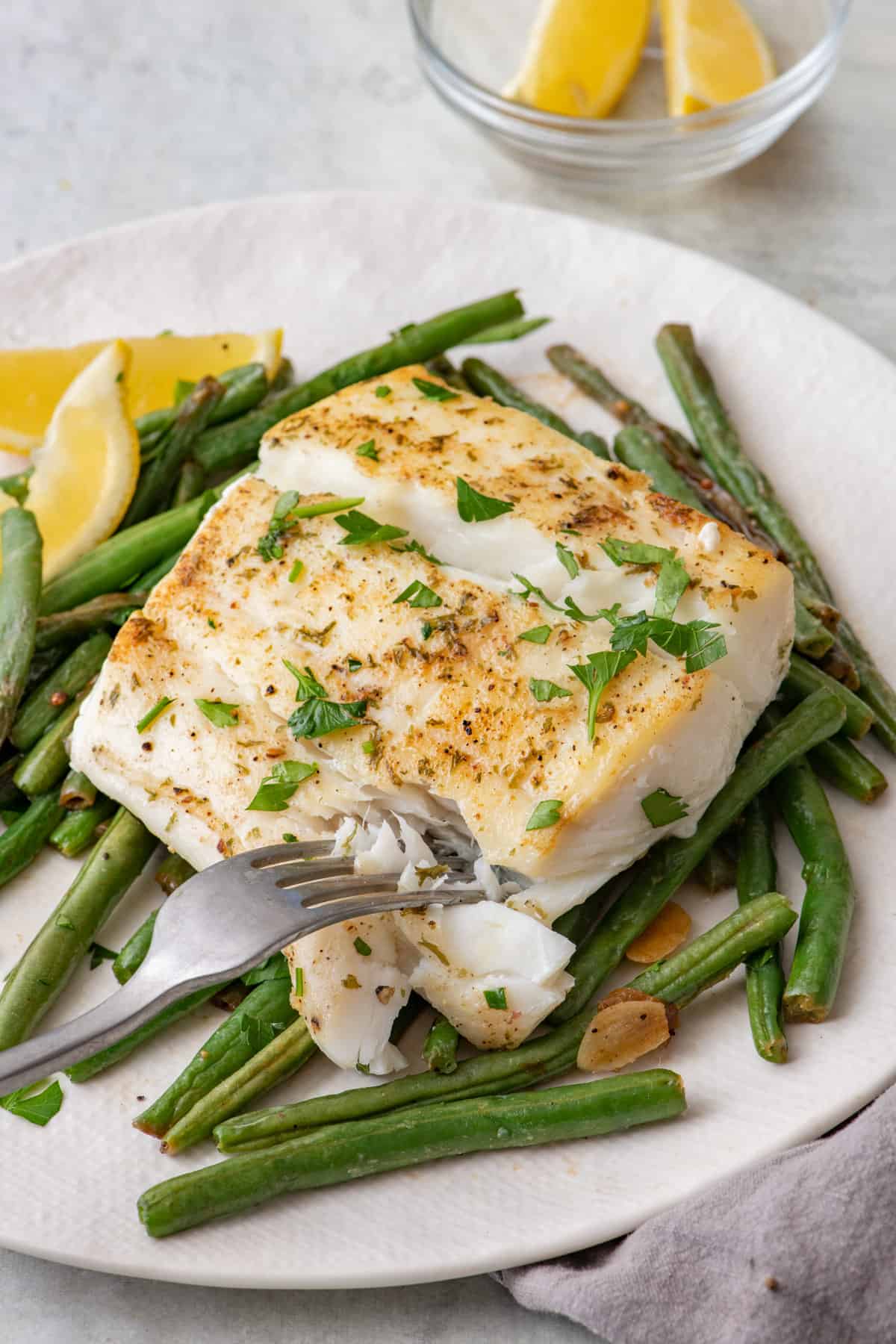 Pan seared halibut fillet on top of green beans garnished with lemon wedges and parsley.