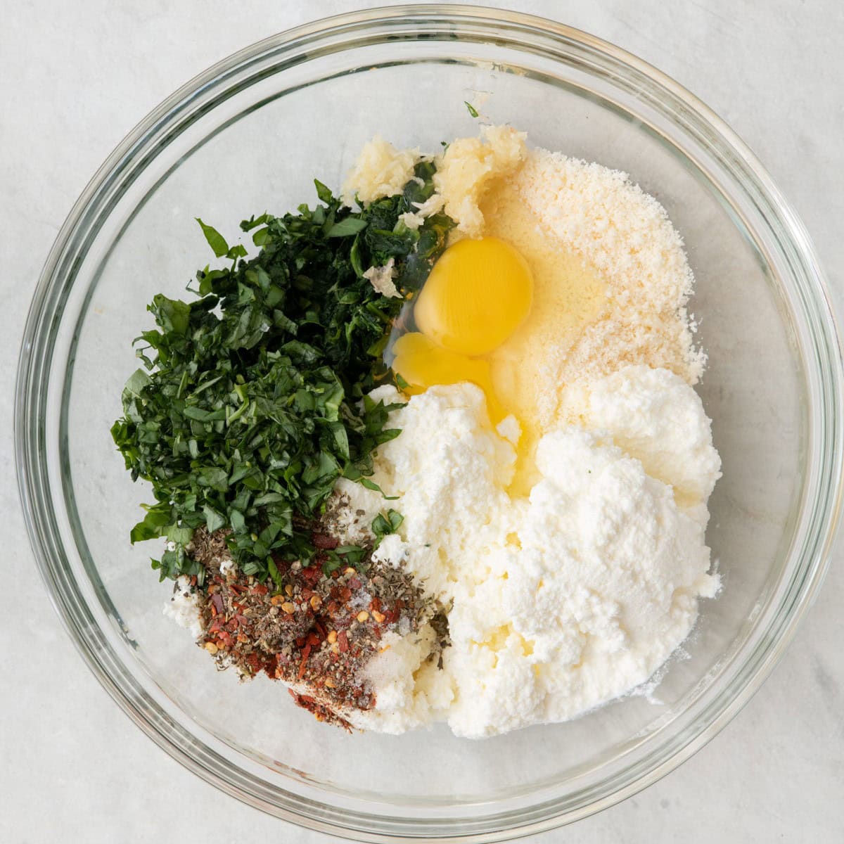 Drained Spinach in a bowl with ricotta, parmesan, egg, garlic, basil, italian seasoning, salt, pepper, and red pepper flakes added, not mixed.