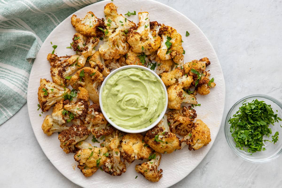 Plate of cooked cauliflower florets with a small bowl of avocado crema in the center and garnished with fresh chopped parsley.