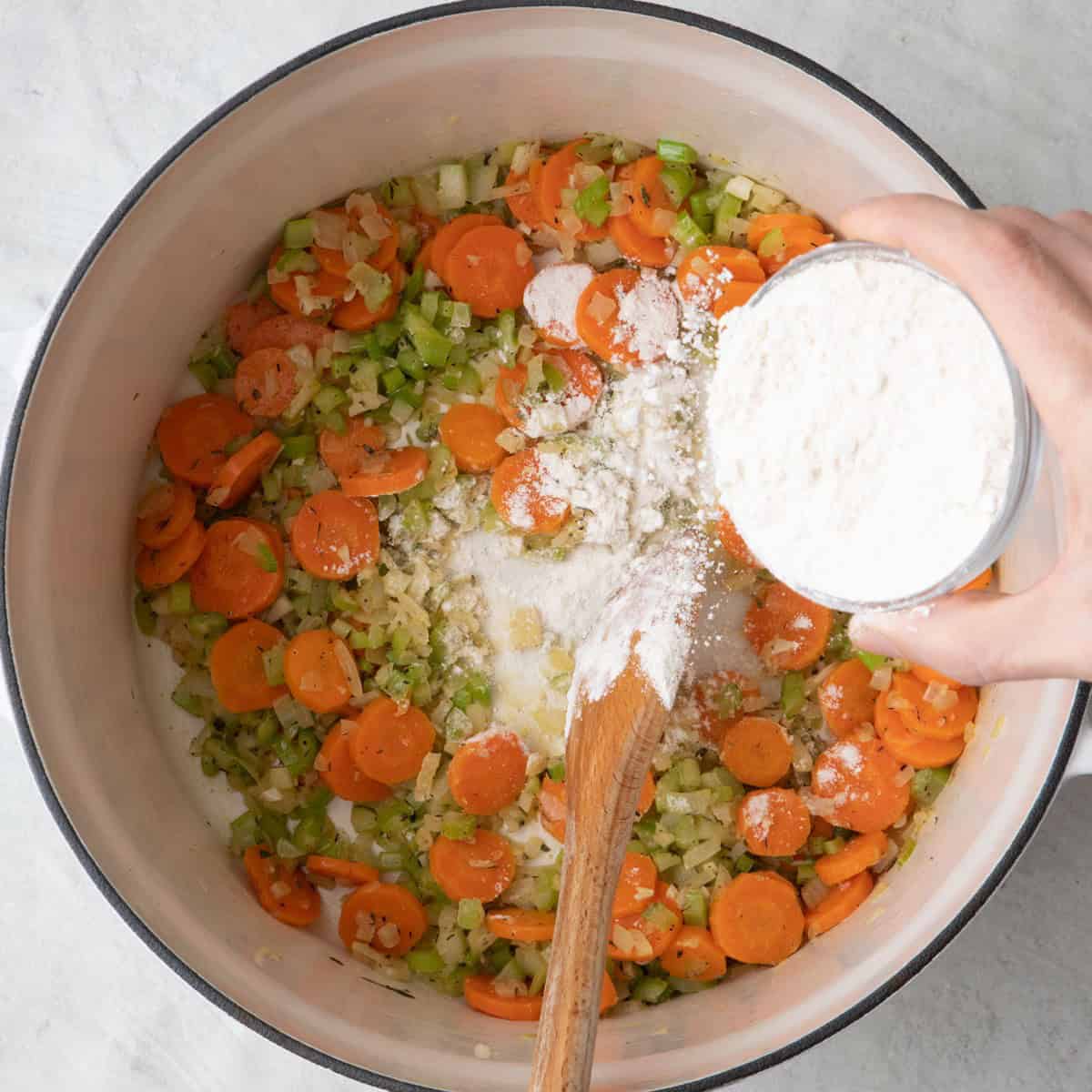 Flour sprinkled over veggies with wooded spoon stirring pot.