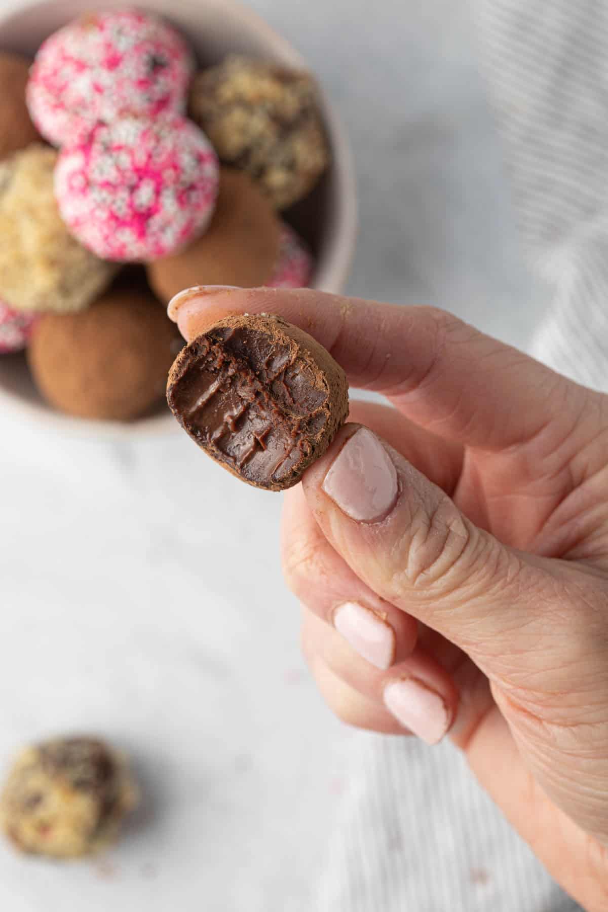 Hand holding one truffle in front of bowl of truffles with a bite removed to show texture inside.