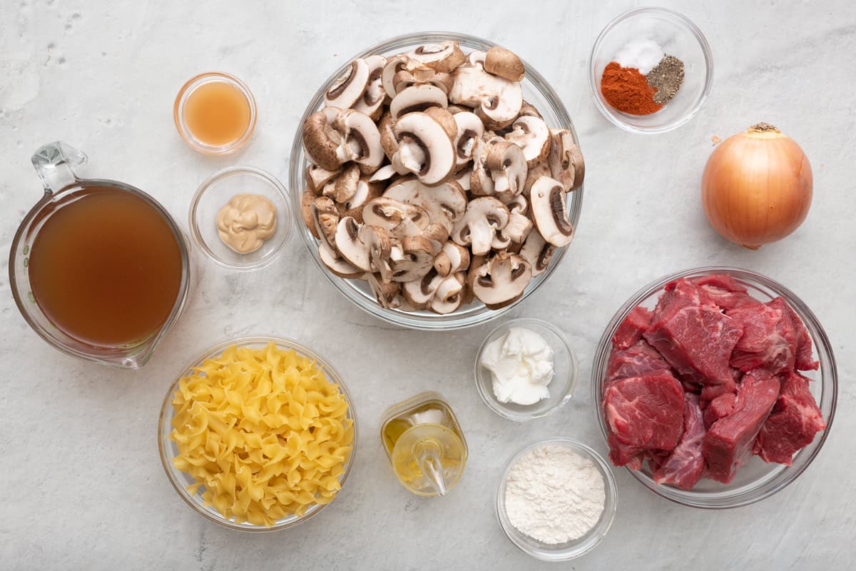 Ingredients for recipe before prepped: broth, apple cider vinegar, dijon mustard, uncooked pasta, sliced mushrooms, oil, greek yogurt, flour, spices, onion, and beef chuck cubes.