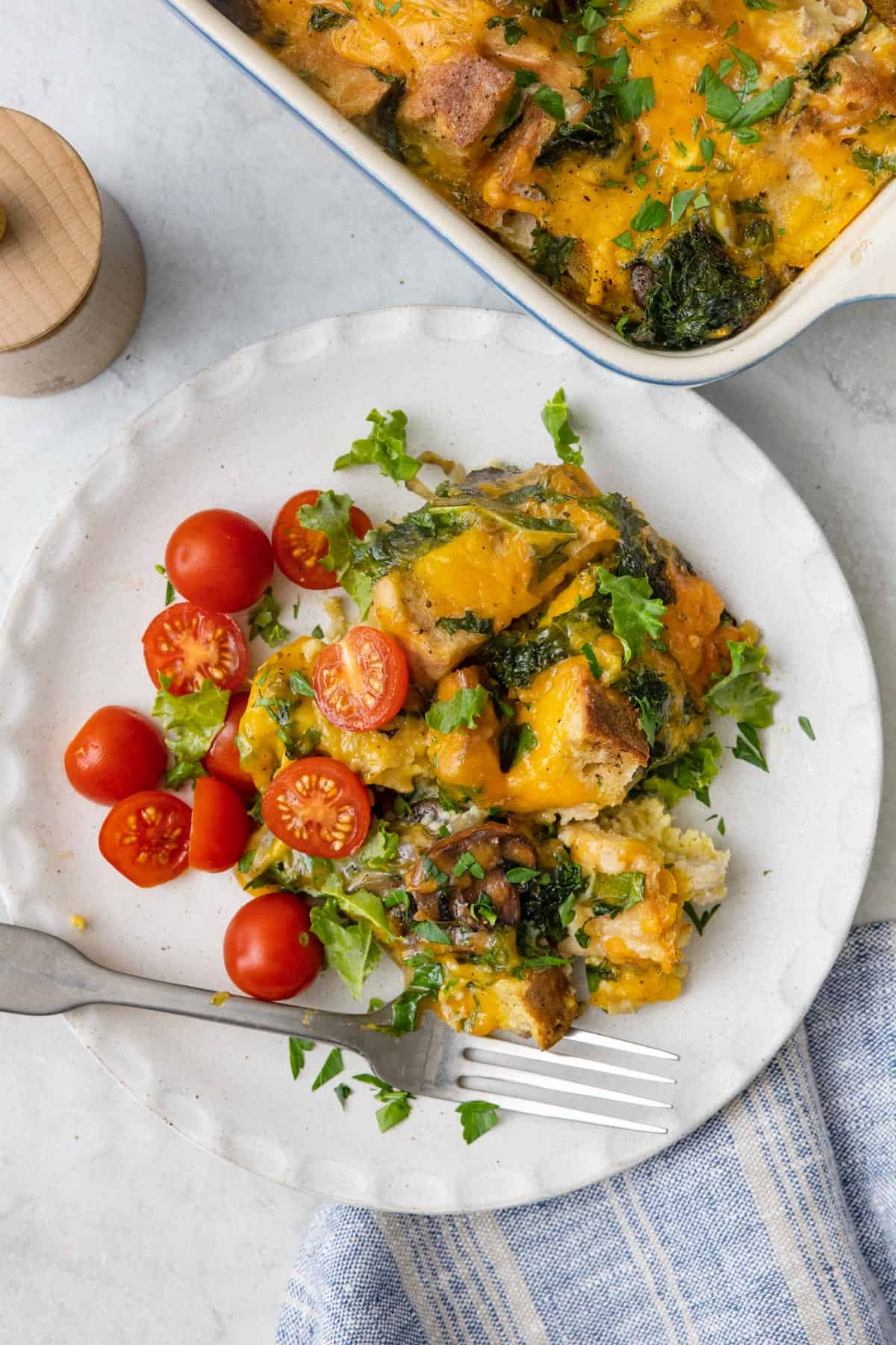 Plate of cheesy breakfast strata with halved cherry tomatoes garnished with fresh herbs and a fork with baking dish of recipe nearby.