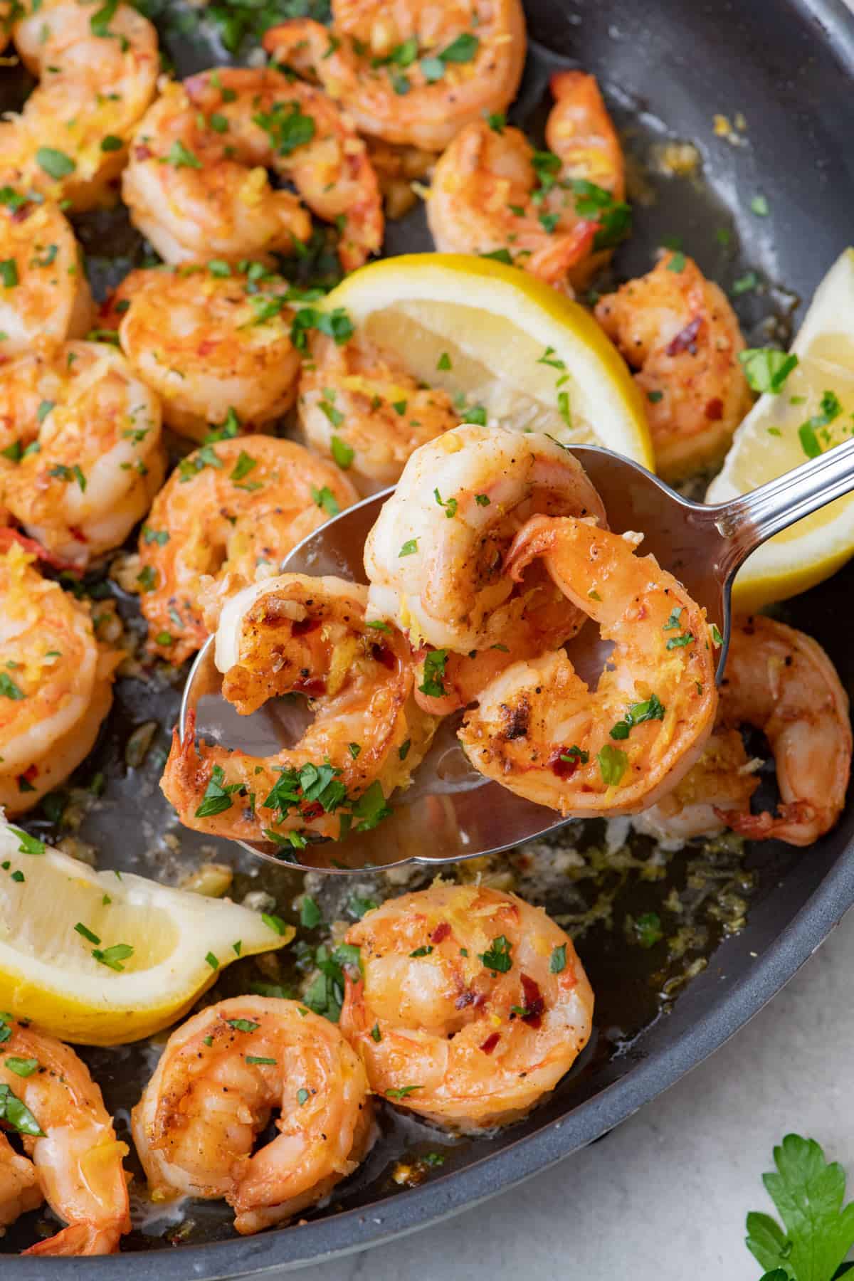 Garlic shrimp in pan with spoon lifting some to show plump shrimp with lemon wedges and parsley.