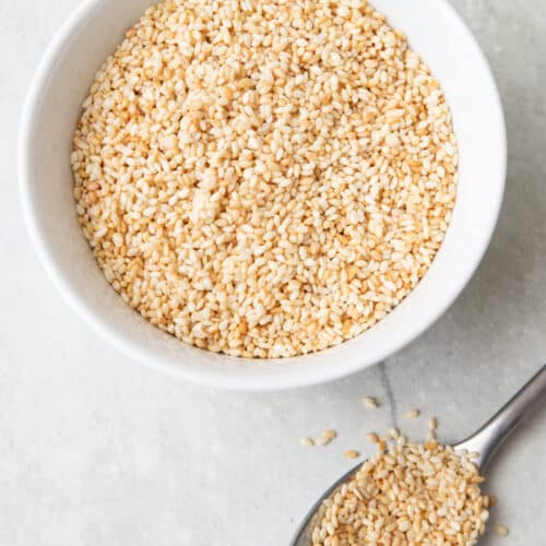 Small serving bowl of toasted seasame seeds with a spoon next to it overflowing with more seeds.