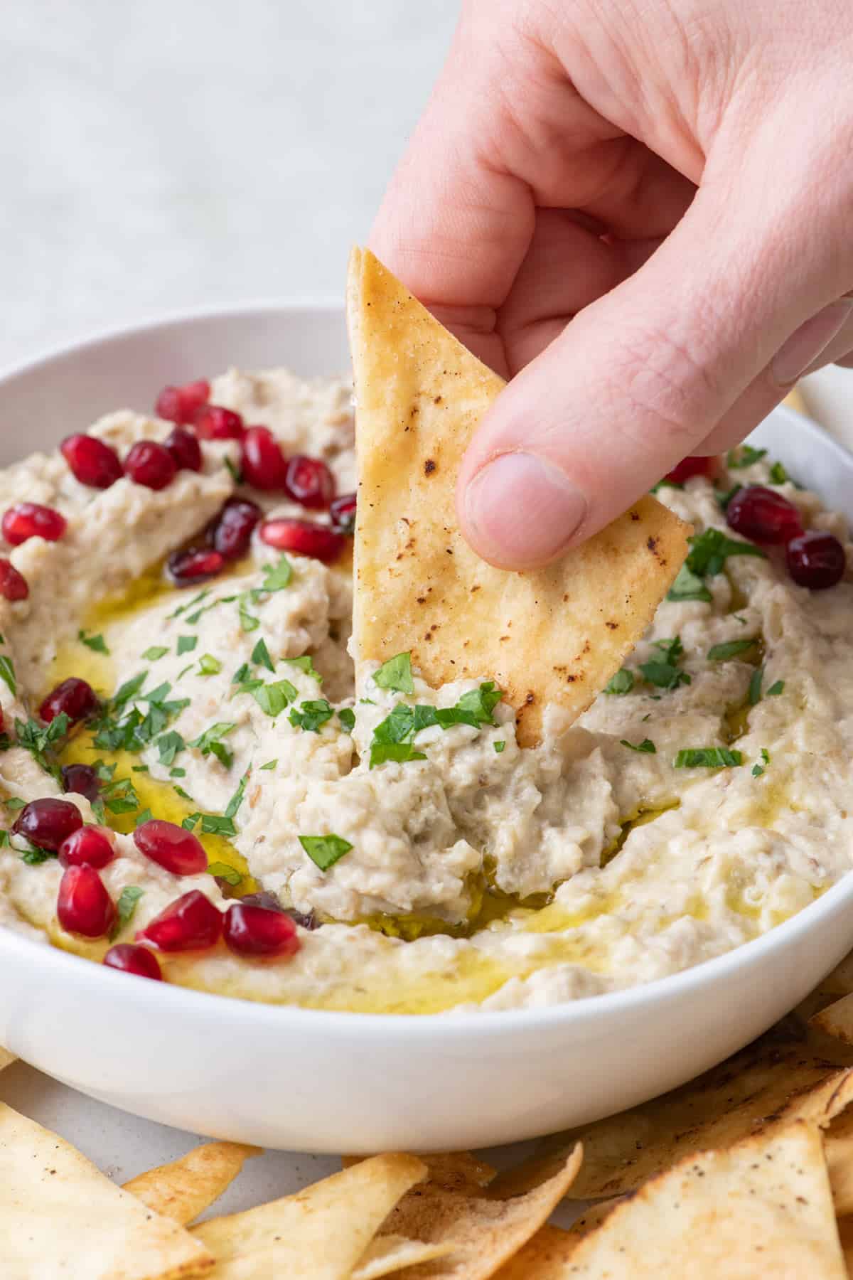 Pita chip being dipped into Baba ghanoush garnished with oil, parsley, and pomegranate seeds.