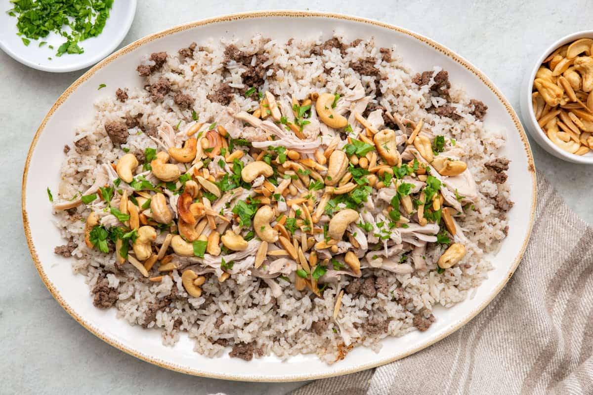 Large oval platter with Lebanese shredded chicken and rice with ground beef crumbles throughout, topped with toasted pinenuts, cashews, and almonds, and garnished with fresh chopped parsley.