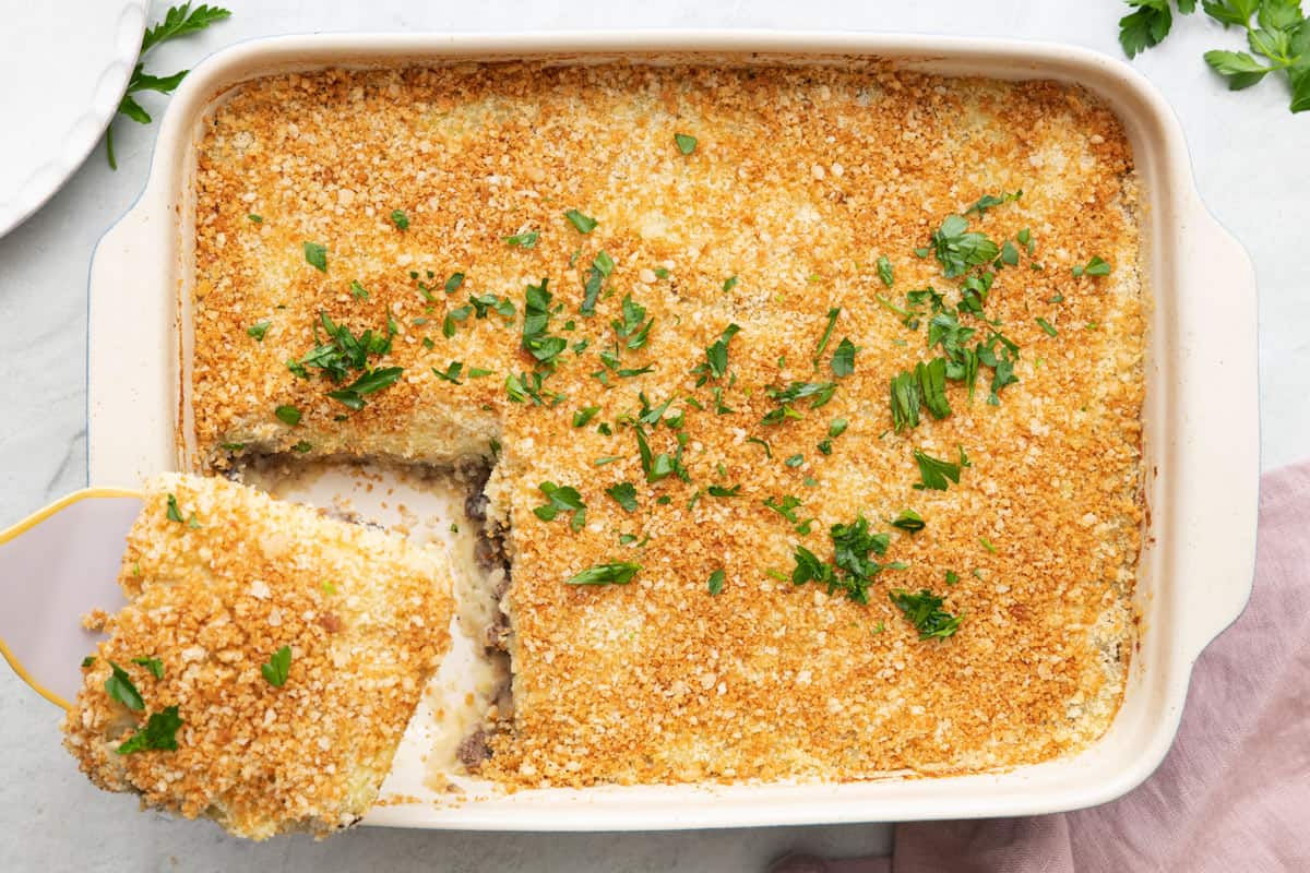 Lebanese potato souffle in a casserole dish with a square serving being removed with a spatula, garnished with fresh parsley.