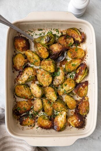 Parmesan crusted brussel sprouts in a baking dish flipped cut side up with crusted cheese with a serving spoon tucked and garnished with fresh parsley and flaky salt.