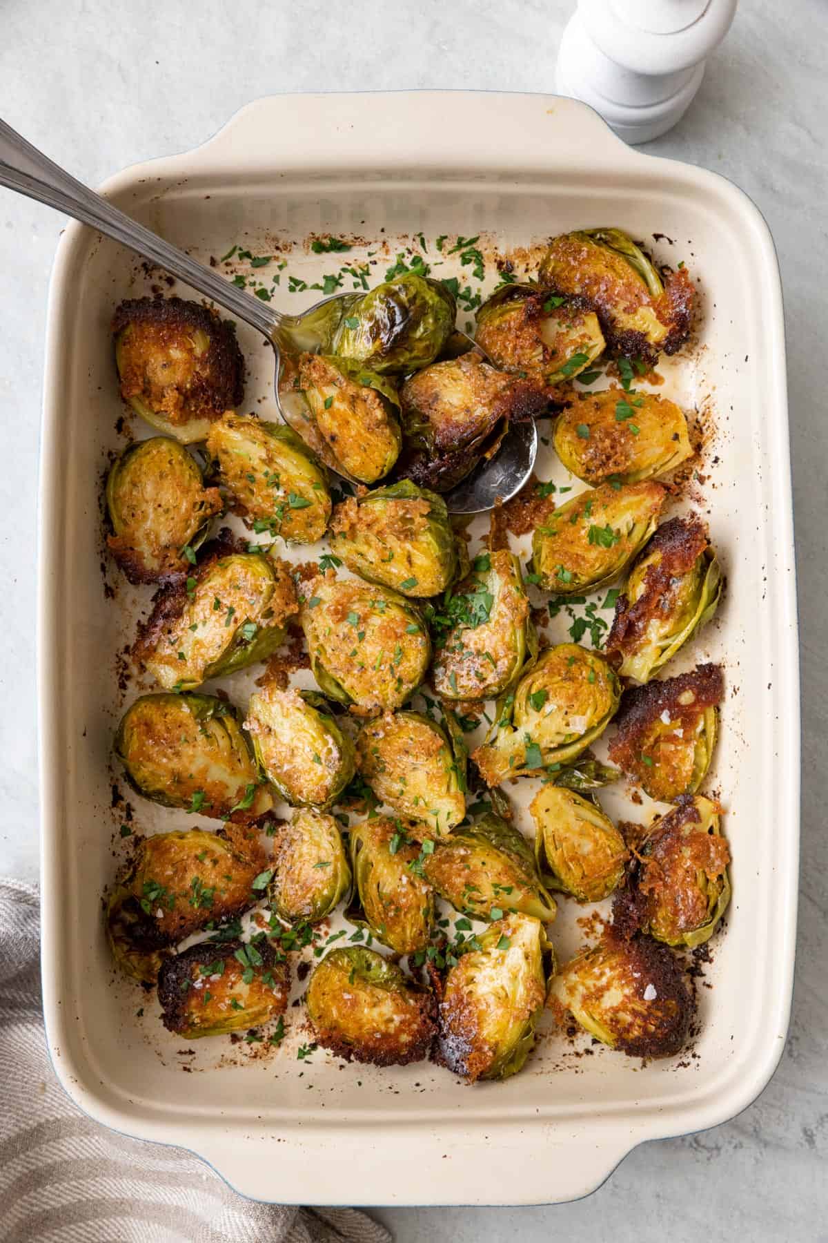Parmesan crusted brussel sprouts in a baking dish flipped cut side up with crusted cheese with a serving spoon tucked and garnished with fresh parsley and flaky salt.