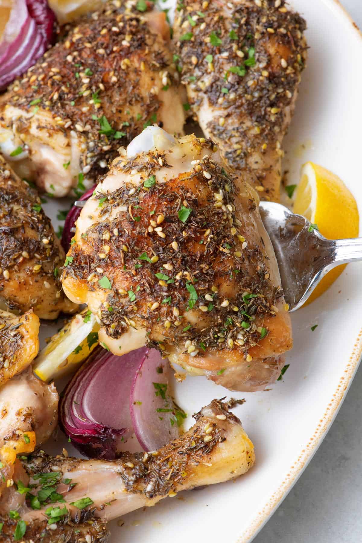 Serving fork lifting up a seasoned chicken thigh from a platter with more chicken pieces.