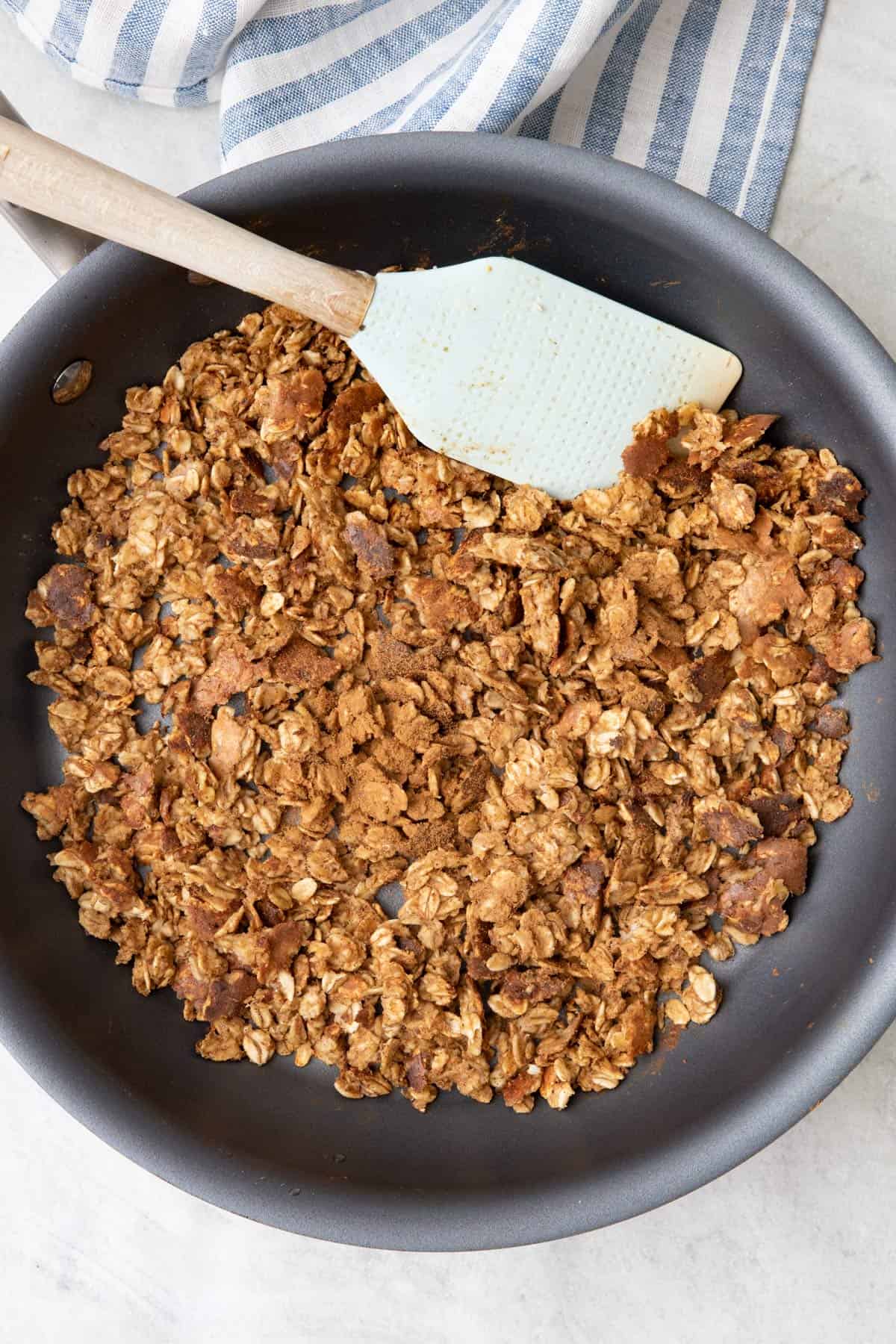 Scrambled oats in pan with spatula set on the side of the pan rim with extra cinnamon sprinkled on top.