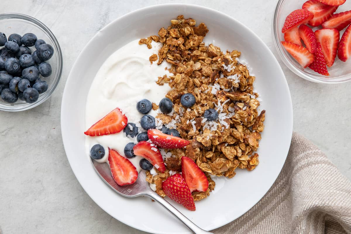 Bowl with scrambled oats, greek yogurt, a few quartered strawberries, blueberries, with coconut sprinkled on top. Small dishes of berries nearby.