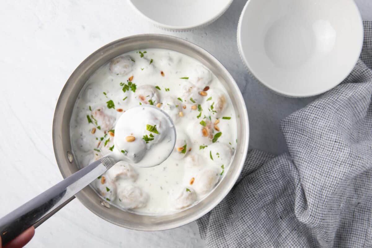 Ladle scooping out a serving of beef filled dumpings from a pot of yogurt sauce with two bowls neaby.
