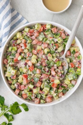 Large white bowl of a chopped vegetable salad tossed in a tahini dressing with a spoon dipped in.