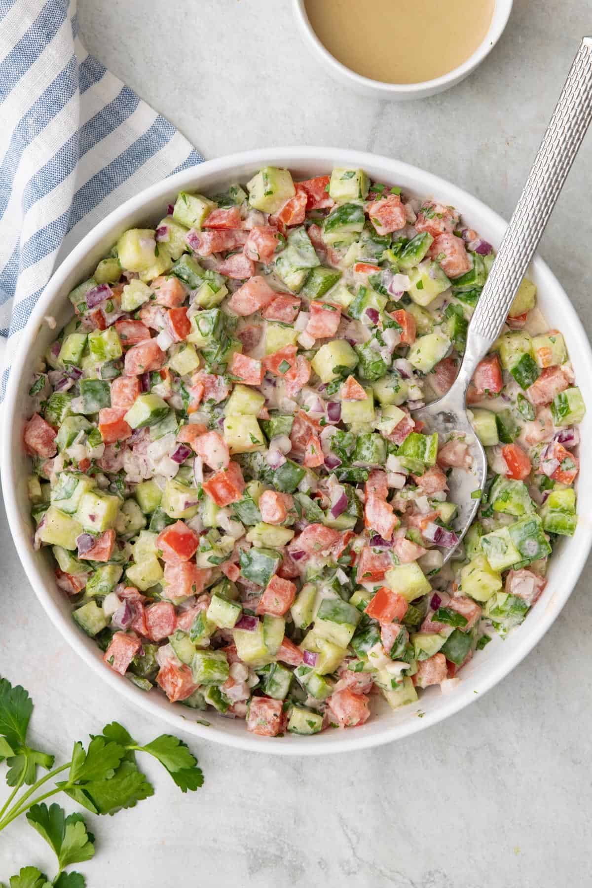 Large white bowl of a chopped vegetable salad tossed in a tahini dressing with a spoon dipped in.