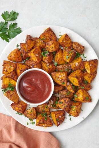 Round plate of crispy air fried seasoned potatoes garnished with flaky salt and fresh herbs, served with a small bowl of ketchup.